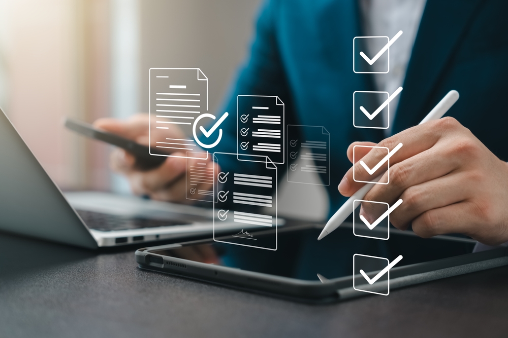 Illustration: ein Mann sitzt am Schreibtisch mit einem Notebook und schreibt auf einem Tablet, sowie Symbole mit abgehakten Kästchen im Vordergrund