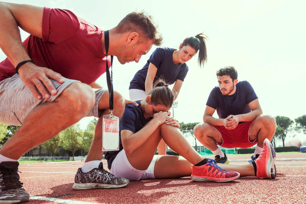Illustration: eine junge Frau in Sportkleidung sitzt auf einer Laufbahn und hält mit den Händen das rechte, verletzte Knie, während sie von zwei jungen Männern und einer Frau sorgenvoll umringt wird