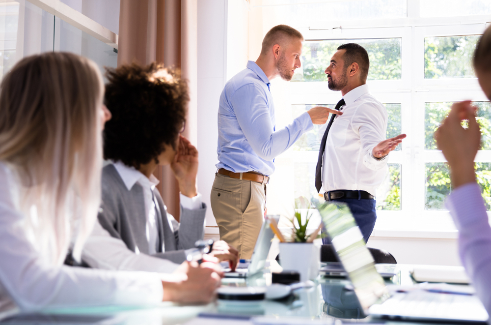 Illustration: bei einer Bürobesprechung sitzen mehrere Personen an einem Tisch im Vordergrund, wobei im Hintergrund zwei Männer stehen und sich mit Drohgebärden streiten