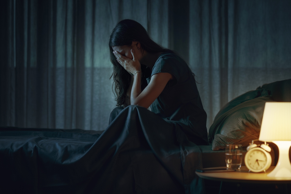 Illustration: eine junge Frau sitzt mit den Händen vor ihrem Gesicht am Bett in einem dunklen Schlafzimmer mit der leuchtenden Nachtischlampe